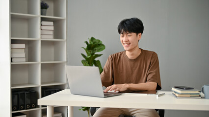 A happy and handsome young Asian man using his laptop in his home office.