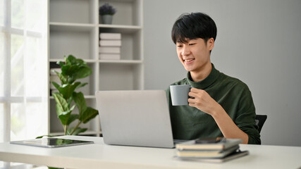 A smart Asian man sipping coffee while working on his tasks on his laptop at his desk