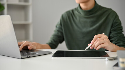 Cropped image of a smart Asian man using his digital tablet and laptop at his desk.