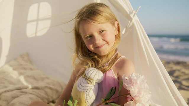 Happy girl in rays of sunset sun. Close up beautiful little girl face. Girl with toy dreaming at ocean beach. Happy face of preschooler looking at RED camera slow mo. Cute smiling child face close up