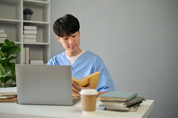 A smart and focused young Asian male medical student in scrubs reading a book at his desk
