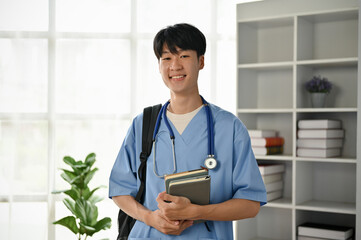 A smiling and kind young Asian male doctor or medical student in scrubs stands in the office.