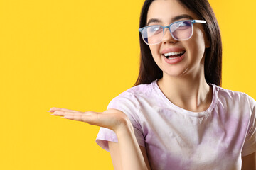 Pretty young woman wearing glasses and showing something on yellow background