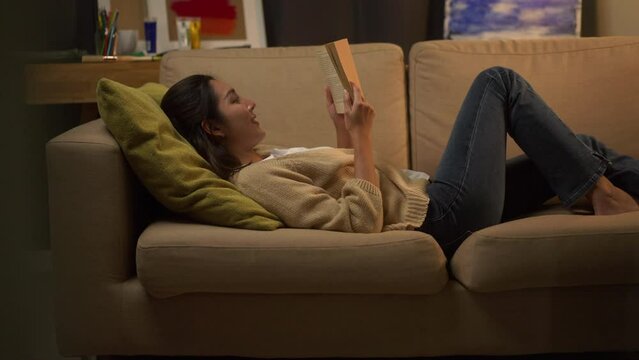 Happy woman opening, reading and relaxing a book on relax time at home. Vacation time of female reading a book on sofa at night. Education concept.