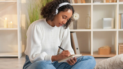 Goal achievement. Home rest. Dream planning. Inspired woman in headphones writing in diary sitting sofa in living room.