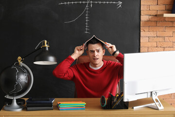 Stressed male teacher with copybook sitting at table in classroom