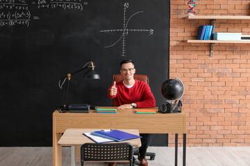 Male teacher showing thumb-up at table in classroom
