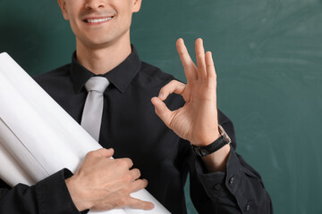 Male teacher with rolls of paper showing OK near chalkboard, closeup