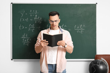 Male Math teacher with book conducting lesson near chalkboard in classroom
