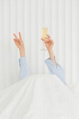 Hand of young woman in a blue pijamas lying in bed under the blanket with champagne glass. Alcohol, celebration and morning concept