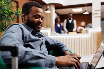 Smiling young African American man entrepreneur using laptop while waiting with suitcase in hotel lobby. Businessman working remotely during business trip, looking at computer screen