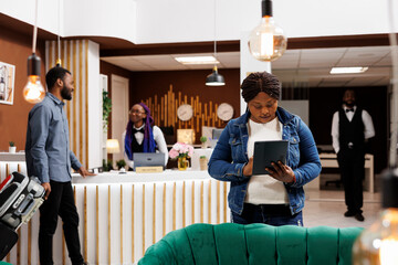 African American woman tourist standing in hotel lobby with digital tablet in hands doing...