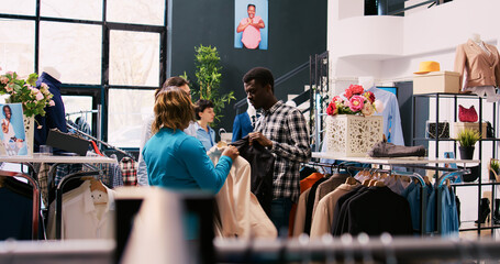 Asian stylish employee helping couple with formal outfit, discussing merchandise fabric in modern boutique. African american customers shopping for fashionable clothes in clothing store