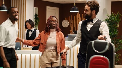 Happy african american tourists tipping helpful bellboy carrying their luggage in resort lobby. Cheerful guests checking out from hotel after handing over room access key card to friendly receptionist - obrazy, fototapety, plakaty