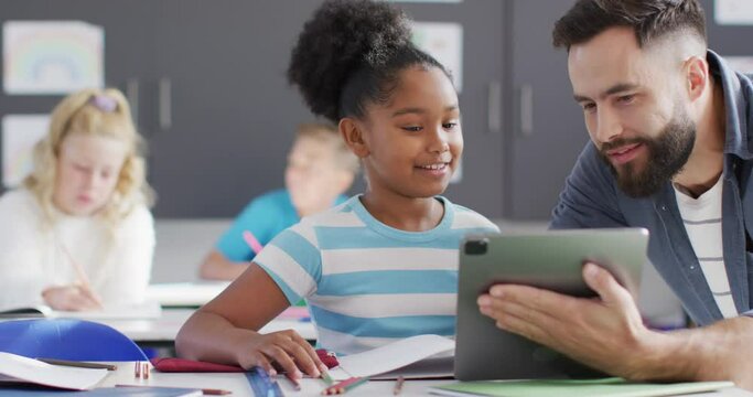 Diverse male teacher and happy schoolchildren using tablet in school classroom