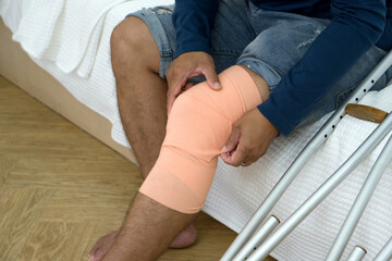 A man with a broken knee sitting on his bed. He leans on crutches, signalling recovery from injury. His bedroom serves as a backdrop, tinged with hints of helplessness yet enduring determination.