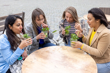 A lively image featuring a multi-ethnic group of female friends savoring refreshing Mojito cocktails on a cafe terrace located in a bustling European city center. The scene encapsulates camaraderie