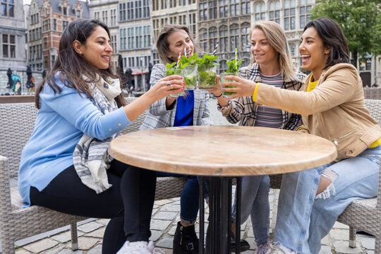 A vibrant photograph depicting a multi-racial group of four female friends sharing a toast with refreshing Mojito cocktails at an outdoor cafe situated in a lively European city center. The image - Powered by Adobe