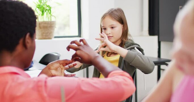 Diverse female teacher and happy schoolchildren at desks learning sign language in school classroom