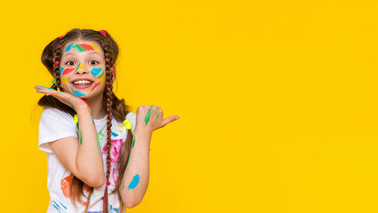 A child smeared with paint. A little girl painted with multicolored paints points to your advertisement. Children's creativity. Yellow isolated background. Copy space. Banner.