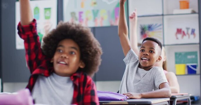 Happy diverse schoolchildren at desks raising hands in school classroom