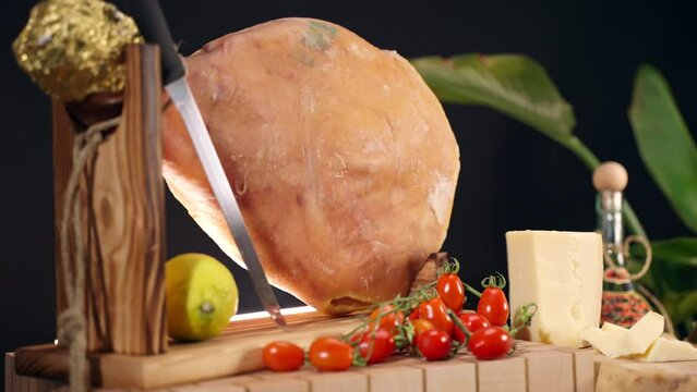 Delicious jamon leg on wooden stand with knife for slicing, lemon and fresh tomatoes against black background, appetising antipasto of Italian cuisine, typical cured prosciutto, meat delicatessen