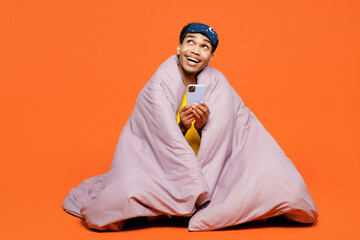 Full body young man wearing pyjamas jam sleep eye mask sit wrapped in duvet rest relax at home use mobile cell phone look aside isolated on plain orange background studio. Good mood night nap concept.