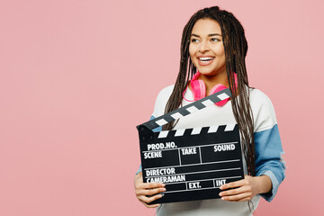 Young woman of African American ethnicity wears white sweatshirt casual clothes hold classic black film making clapperboard look aside isolated on plain pastel light pink background studio portrait.