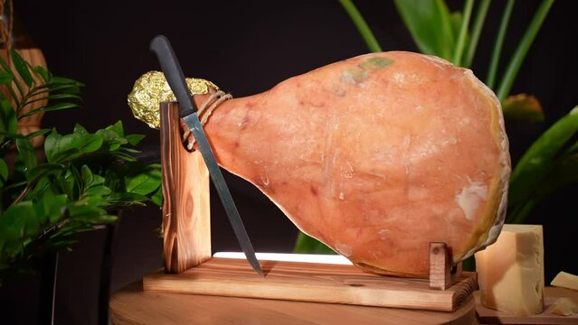 Cured parma ham on wooden stand with knife for slicing against black background, dry salted prosciutto leg is an appetising antipasto of mediterranean cuisine, traditional Italian and Spanish meal