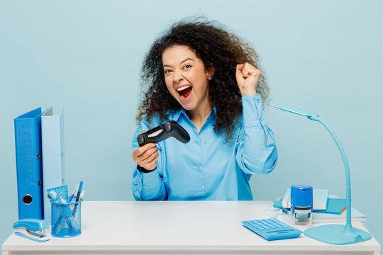 Young successful employee business woman wearing casual shirt sit work at white office desk hold in hand play pc game with joystick console do winner gesture isolated on plain pastel blue background.