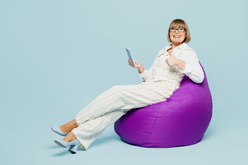 Full body employee business woman 50s wear white classic suit glasses formal clothes sit in bag chair use mobile cell phone show thumb up isolated on plain blue background. Achievement career concept.