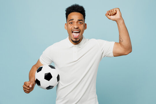 Young happy man fan wears basic t-shirt cheer up support football sport team hold in hand soccer ball watch tv live stream do winner gesture clenching fists isolated on plain blue background studio.