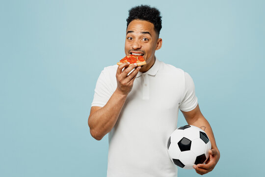 Young excited happy cheerful fun man fan wear basic t-shirt cheer up support football sport team hold in hand soccer ball eat pizza watch tv live stream isolated on plain pastel blue color background.
