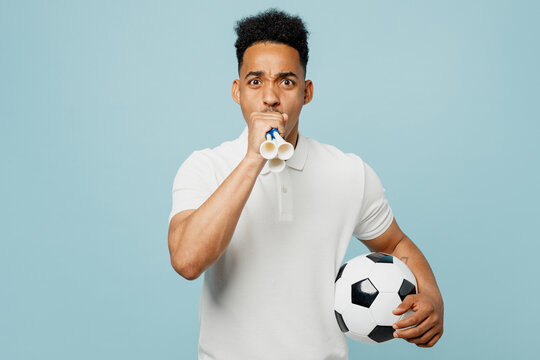 Young happy fun man fan wear basic t-shirt cheer up support football sport team hold in hand soccer ball blowing pipe look camera watch tv live stream isolated on plain pastel blue color background