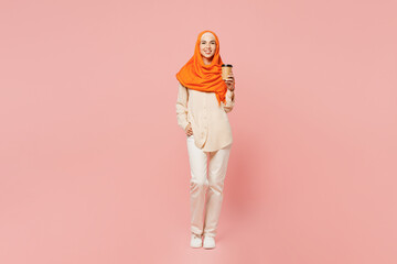 Full body young arabian asian muslim woman wears orange abaya hijab hold takeaway delivery craft paper cup coffee to go isolated on plain pink background. Uae middle eastern islam religious concept.