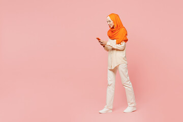 Fototapeta na wymiar Full body side view young arabian asian muslim woman wear orange abaya hijab hold in hand use mobile cell phone walk go isolated on plain pink background. Uae middle eastern islam religious concept.