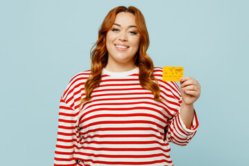 Young smiling happy chubby overweight woman wear striped red shirt casual clothes hold in hand mock up of credit bank card isolated on plain pastel blue background studio portrait. Lifestyle concept.