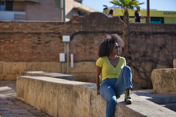 Fototapeta na wymiar Young, beautiful, black woman with afro hair, wearing yellow t-shirt, jeans and sunglasses, sitting on a stone wall, relaxed, calm and sunbathing. Vacation concept, travel, current, modern.