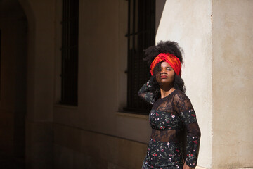 Fototapeta na wymiar Young, beautiful and black woman with afro hair, with a floral dress and scarf in her hair, leaning on a wall receiving the sun's rays. Concept beauty, ethnicity, fashion, portraits.