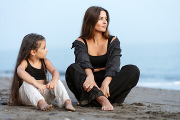 Fototapeta na wymiar Romantic young mother and little daughter hugging, sitting at the beach and looking away