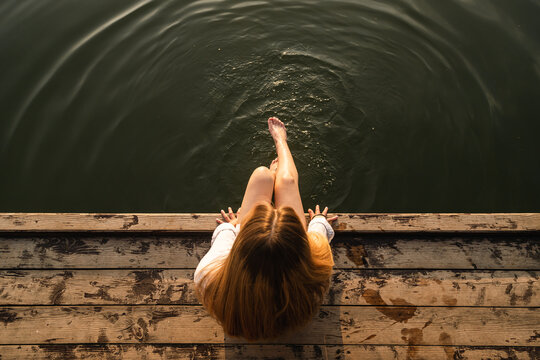 Woman relaxes by the lake sitting on the edge of a wooden jetty, swing feet near the water surface, top view.