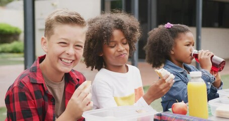 Video of three diverse schoolchildren eating packed lunch and laughing in schoolyard