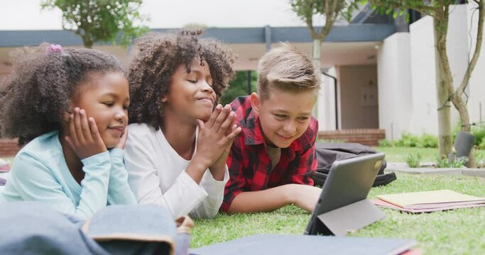 Video of three happy diverse schoolchildren watching tablet lying in schoolyard, copy space