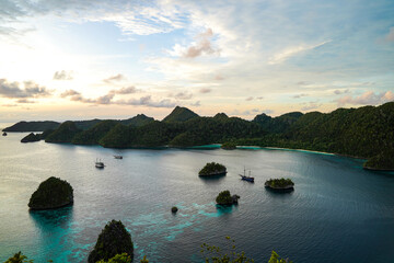 A view of the blue lagoon with a sunset sky, clear sea, some sailing ships and limestone islands in remote archipelago. View from the top of the Wayag Islands, Raja Ampat, West Papua, Indonesia.