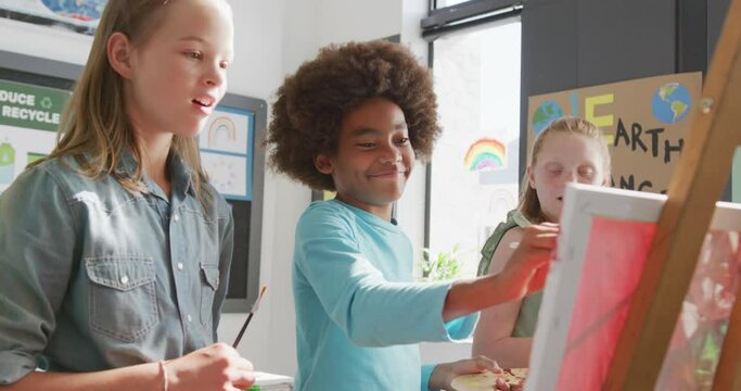 Video of diverse, happy schoolchildren standing at easels painting, talking in art class, copy space