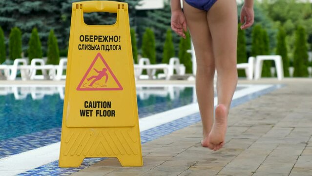 The inscription in Ukrainian carefully wet floor. Girl child walks past a yellow yellow warning sign that the floor near the pool is slippery