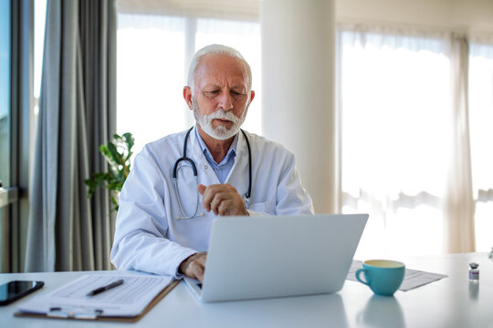 Serious mature doctor using laptop and sitting at desk. Senior professional medic physician wearing white coat and stethoscope working on computer at workplace.