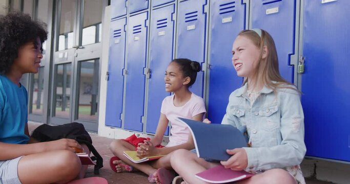 Video of three happy, diverse schoolgirls talking, sitting by lockers in school corridor, copy space