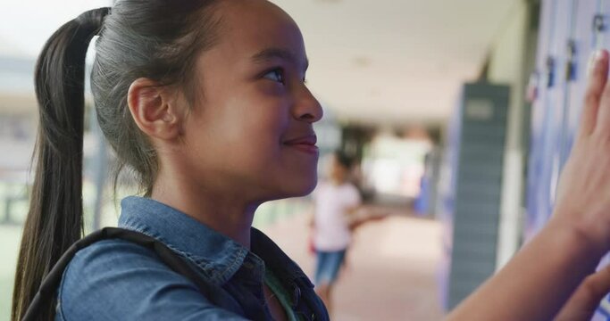Video portrait of happy biracial schoolgirl using locker, smiling in school corridor, copy space