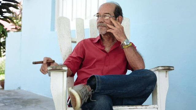 Senior Pensive Cuban Man smoking a cigar sitting at home porch looking away